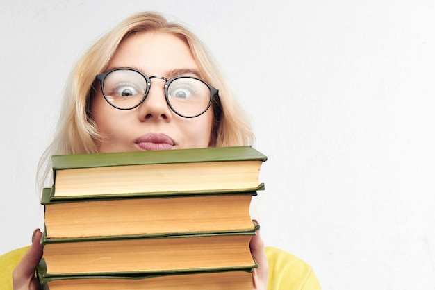 Funny clever girl looks out of a pile of books. Blonde nerd in round glasses on a white background. Educational concept