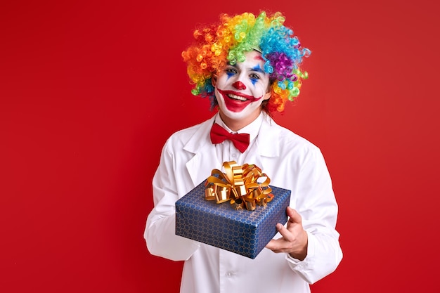 Забавный клоун с подарочной коробкой на красном фоне, молодой арлекин смотрит в камеру
