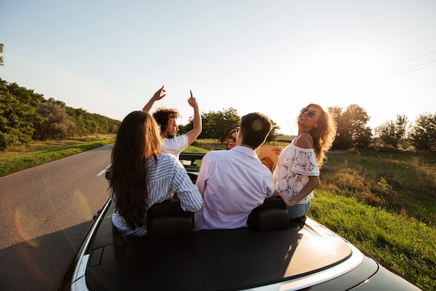 Веселая компания счастливых молодых девушек и парней сидит на дороге черного кабриолета в солнечный день. .