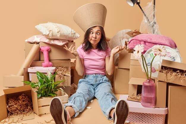 Funny confused asian woman crosses eyes shrugs shoulders has
lampshade on head dressed in t shirt and jeans surrounded by stacks
of cardboard boxes going to relocate to new apartment beige
wall