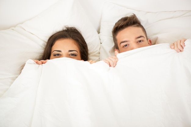 Funny couple in bed looking and peeking over sheets smiling.