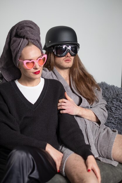 Funny couple in love sitting on sofa towel and helmet on head glasses with hearts and motorcycle goggles on eyes man has bare legs