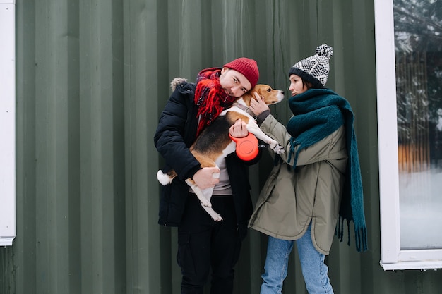 Funny couple trying to pose for a photo with their beagle outdoors at winter. Man is hugging the dog, woman is calming her down. In front of corrugated sheet house.