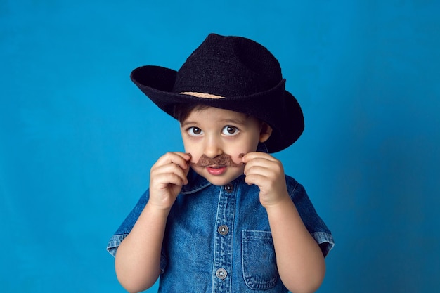 Funny cowboy boy with a mustache and a hat stands in the studio on a blue background