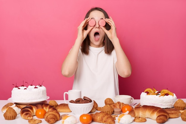 Funny crazy brown haired woman in white t shirt sitting at table with cinfectionery isolated over pink background covering eyes with cookies keeps mouth widely opened