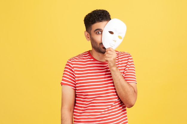 Funny crazy man with beard in striped t-shirt peeping out white face mask, hiding his real feelings, pretending to be another person. Indoor studio shot isolated on yellow background