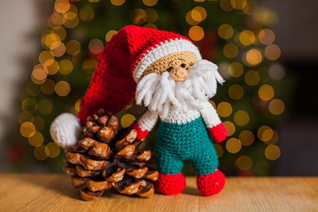 The funny crochet toy gnome in santa hat is standing on a wooden table with a cone near