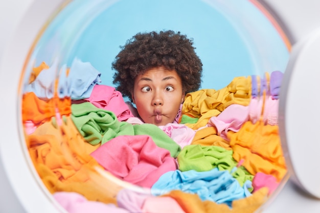 Funny curly Afro American woman housekeeper crosses eyes makes fish lips poses with funny grimace drowned in heap of multicolored laundry has busy day washes clothes involved in housekeeping