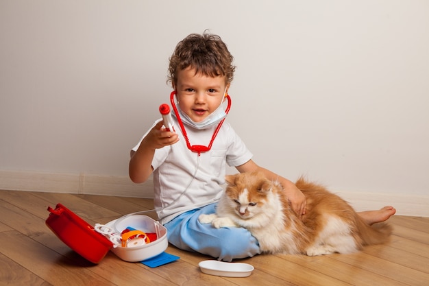 Funny curly kid in a medical mask and glasses with a stethoscope on his neck plays a doctor with a cat.