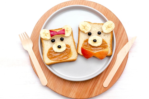 웃긴 귀여운 곰돌이는 땅콩 버터 바나나와 사과를 곁들인 샌드위치 토스트 빵에 직면해 있습니다. 어린이 아기의 달콤한 디저트 건강한 아침 식사 점심 음식 예술 접시흰색 배경 닫기 uptop 보기