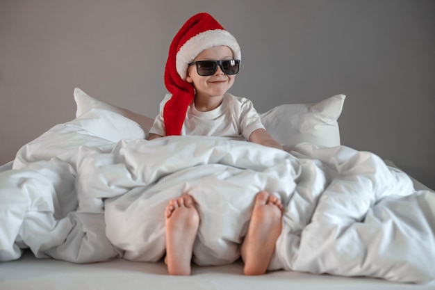 Funny cute boy is sitting on the bed in sunglasses sipping in a Santa hat