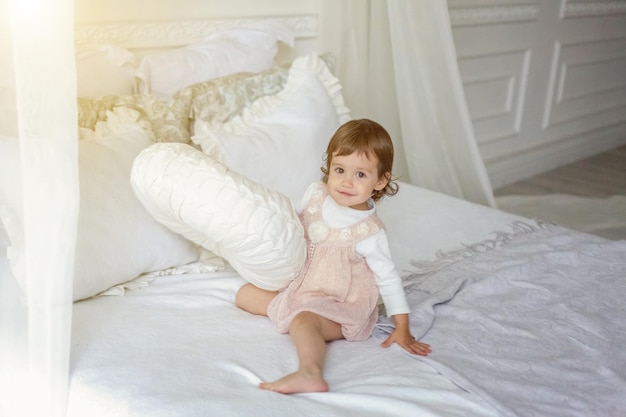 Funny and cute brunette little smiling girl playing jumping on bed in light bedroom. White interior with big bed. Childhood, preschool, youth, relax concept