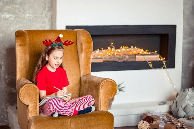 Funny cute caucasian girl lefthanded writes a letter to Santa Claus near the Christmas tree