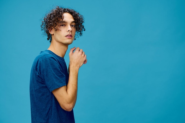 Funny cute curly tanned Caucasian guy in basic tshirt folds hands looks at camera posing isolated on over blue background Lifestyle and Emotions concept Good offer with copy space for ad