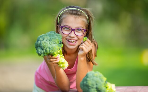 Funny cute girl holding in hands red cabbage and broccoli. Blurred background in garden. Pre-teen young girl with glasses and teeth braces.