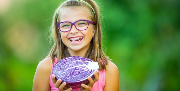 Funny cute girl holding in hands red cabbage and broccoli. Blurred background in garden. Pre-teen young girl with glasses and teeth braces.