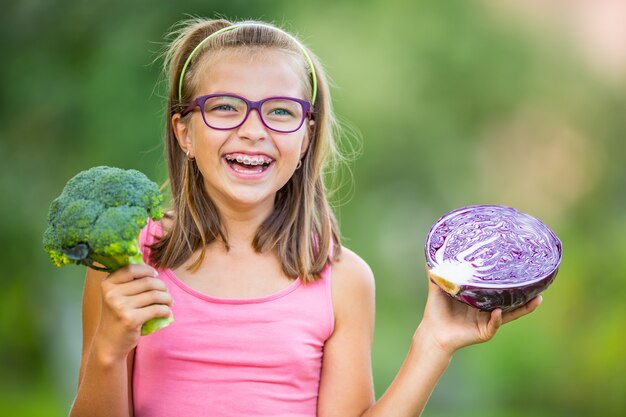 Funny cute girl holding in hands red cabbage and broccoli. Blurred background in garden. Pre-teen young girl with glasses and teeth braces.