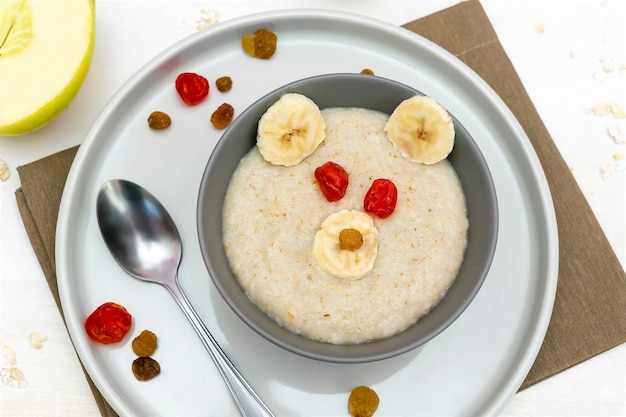 Funny cute kids childrens baby's healthy breakfast lunch oatmeal porridge in bowl look like bear face decorated with apple banana dried berry fruits dessert food art on white wooden table