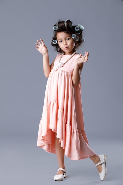 Funny cute little child girl in pink dress and hair curlers posing on gray background. Human emotions and facial expression