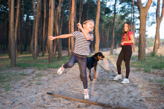 暖かい晴れた夏の明るい朝に密な松林の中で彼女の顔に広い笑顔で大きな古い乾いた枝を飛び越えて面白いかわいい女の子、そして犬と一緒の妹が背景に立っています