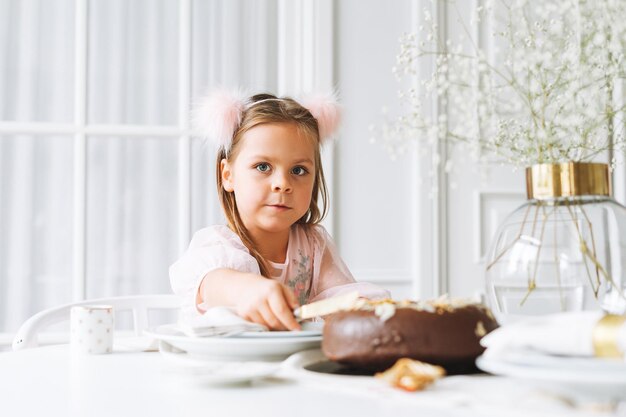 Photo funny cute little girl with long hair in light pink dress with chocolate cake in hands on festive table in bright living room at the home christmas time birthday girl