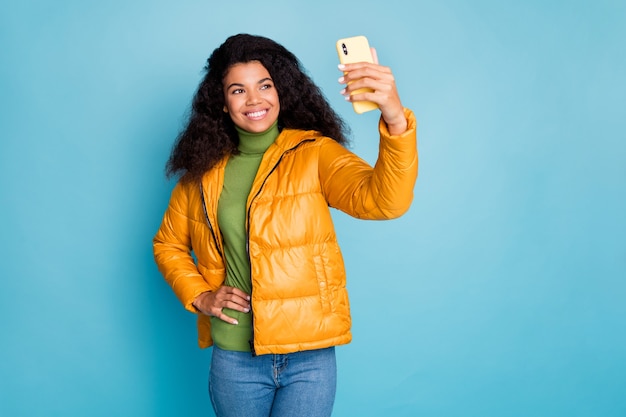 電話を保持している面白い暗い肌の巻き毛の女性selfies現代の流行のヒップスターを着用黄色の秋のオーバーコートジーンズ緑のセーター孤立した青い色の壁