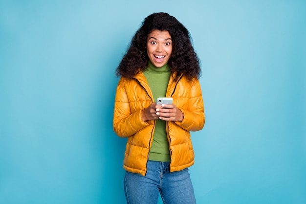 funny dark skin lady open mouth holding telephone hands enjoy new blog followers wear yellow spring overcoat jeans sweater isolated blue color wall