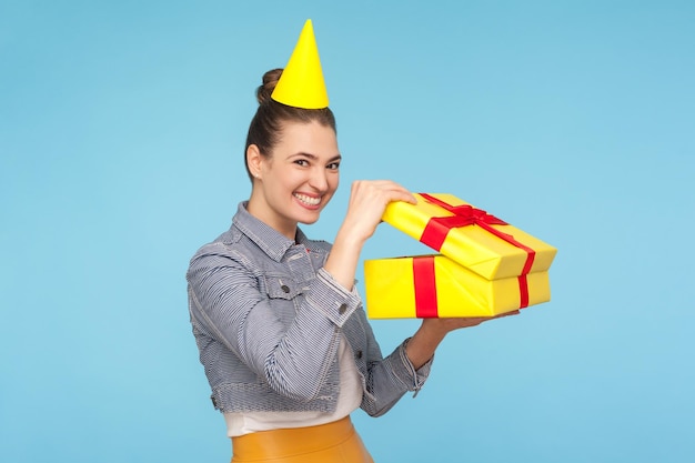 Funny delighted jubilant woman with party cone opening gift box and grinning at camera unpacking present with expression of extreme joy happiness indoor studio shot isolated on blue background