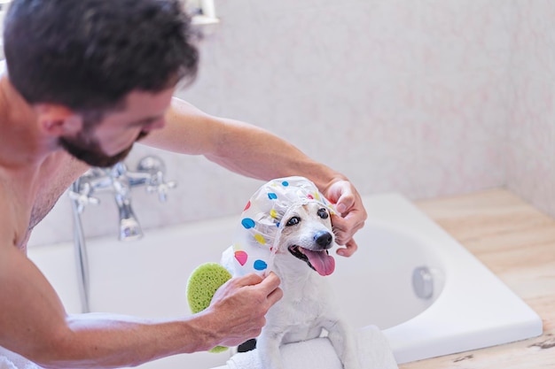 Смешная собака в шапочке для душа развлекается с мыльными пузырями в ванной с хозяином