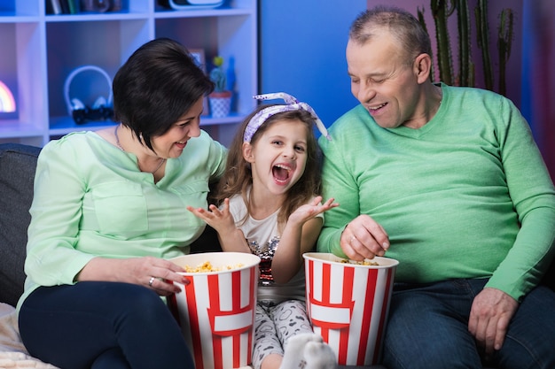 Забавная старшая семейка и внучка маленького ребенка сидят на диване и смотрят телевизор, съедая попкорн