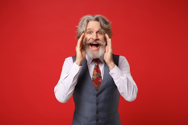 Funny elderly gray-haired mustache bearded man in classic shirt vest colorful tie posing isolated on red background. People lifestyle concept. Mock up copy space. Scream with hand gesture near mouth.