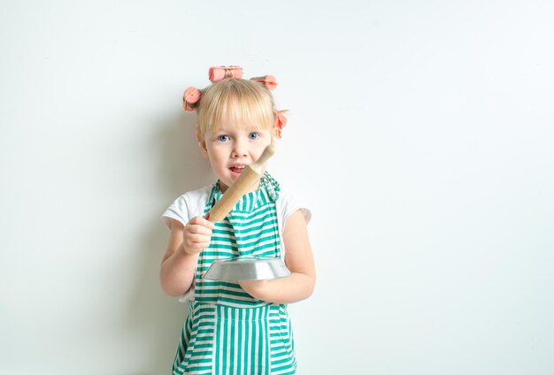 funny emotional child in the role of an angry wife in curlers with a frying pan in a kitchen apron