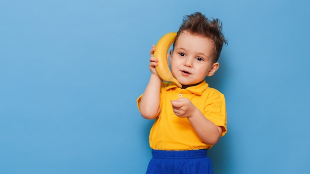 バナナと変な顔。黄色いTシャツを着た男の子が笑顔でバナナを持っています。面白い表現。栄養の概念。新鮮なバナナ。イエロースタイル。青い背景