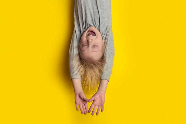 Funny fair-haired boy hanging upside down with arms outstretched and closed eyes. portrait of child on bright yellow background