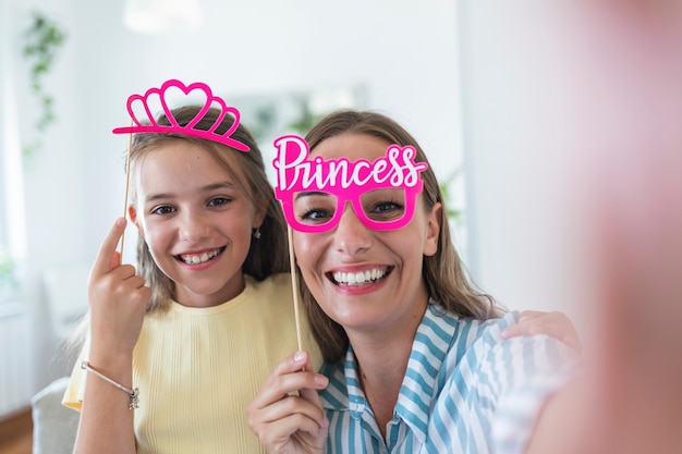 Funny family on a background of bright wall. Mother and her daughter girl with a paper accessories. Mom and child are holding paper crown on stick.