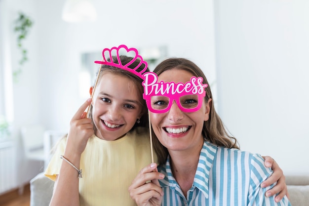 Photo funny family on a background of bright wall. mother and her daughter girl with a paper accessories. mom and child are holding paper crown on stick.