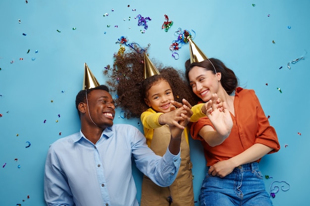 Funny family in caps celebrate birthday. Pretty little girl and her parents, event celebration, balloons and confetti decoration