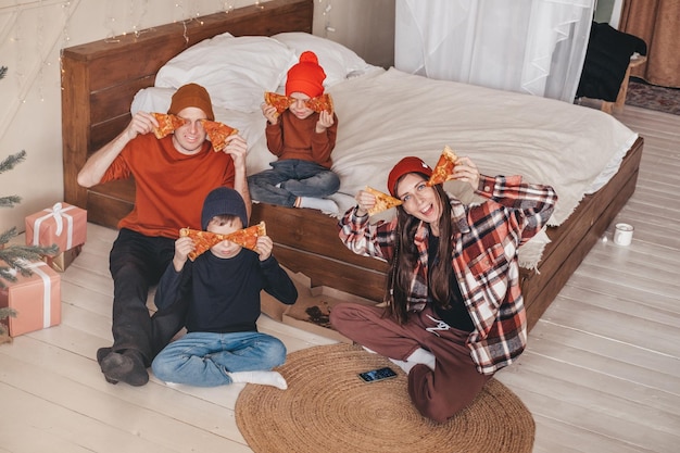 Funny family photo with two children eating pizza in bed happy family moment on the weekend A family with two children is eating an ordered pizza from a box in the bedroom Boho style bedroom