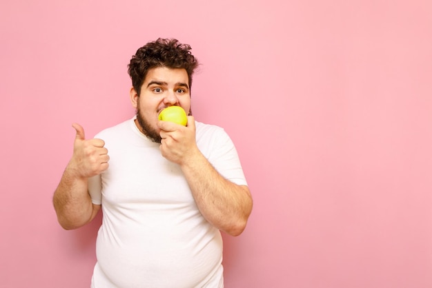 Funny fat guy in a white Tshirt stands on a pink background eats an apple and shows his thumb up