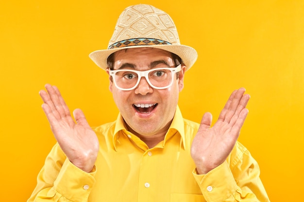 Funny fat man tourist in hat and glasses with surprised face spreads arms on yellow studio background