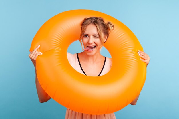 Funny female with two hair buns in dress holding orange rubber ring having rest on resort looking at camera and showing tongue out Indoor studio shot isolated on blue background