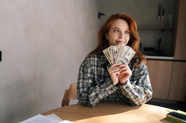Фото Забавная обманчивая молодая женщина подсчитывает деньги в долларах банкноты сидит на кухонном столе дома богатый