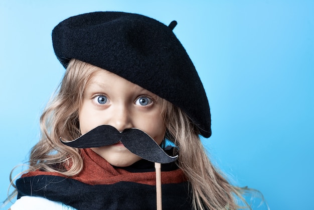 Funny girl in black beret, scarf and mustache on a stick on blue