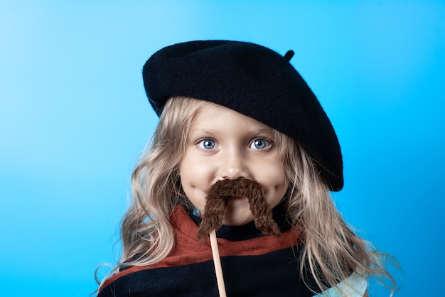 Funny girl in black beret, scarf and mustache on a stick on blue 