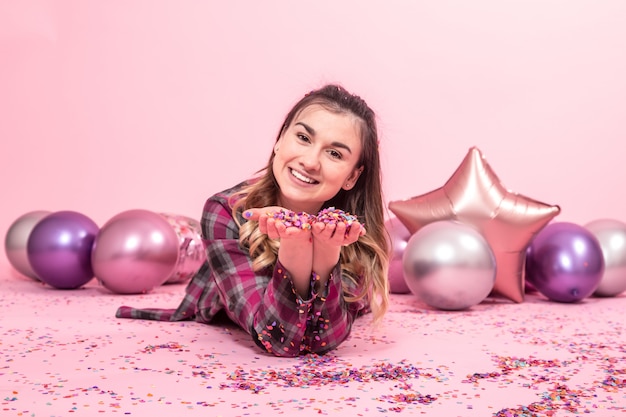 Photo funny girl blows confetti in her hands. color balloons on a pink wall