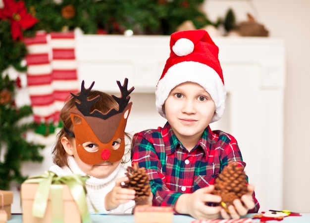 クリスマスの鹿のマスクと松ぼっくりを保持しているサンタの帽子の面白い女の子と男の子