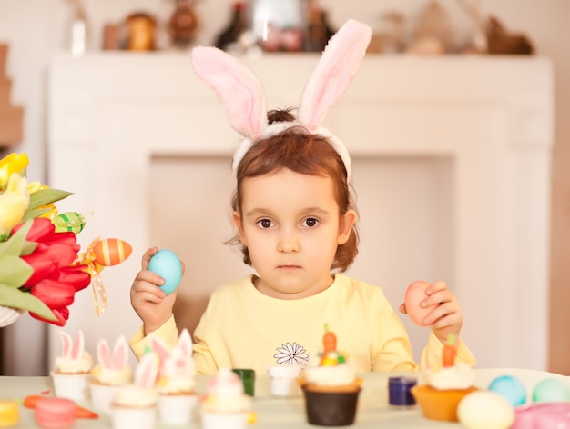 Funny girl child wearing bunny ears and with Easter egg in hand at home.