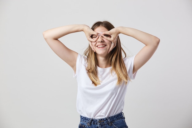 白いTシャツとジーンズに身を包んだおかしな女の子は彼女の目に彼女の手で眼鏡を作ります