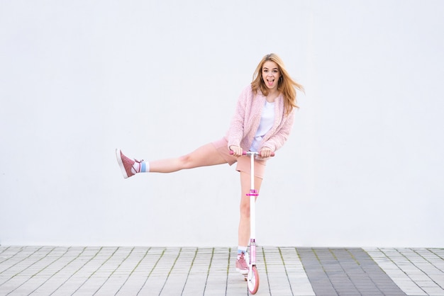 A funny girl in pink clothes goes on a pink kick scooter on the background of a white wall and rejoices.