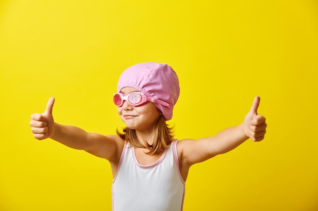 Funny girl in swimsuit stands on yellow background shows thumbs up wearing swimming cap and glasses